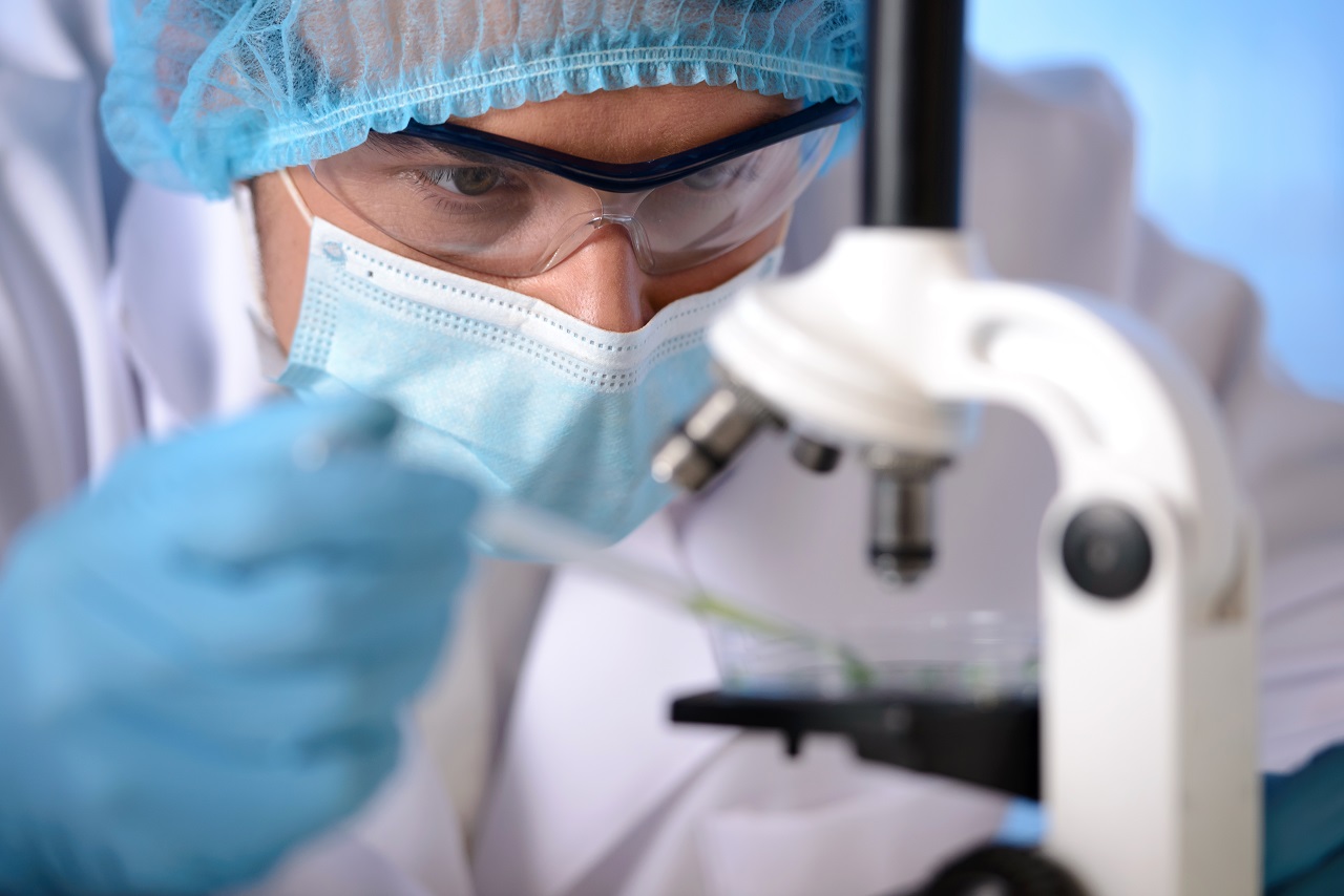 A scientist in a protective mask, glasses, and hair cap dropping a substance to examine