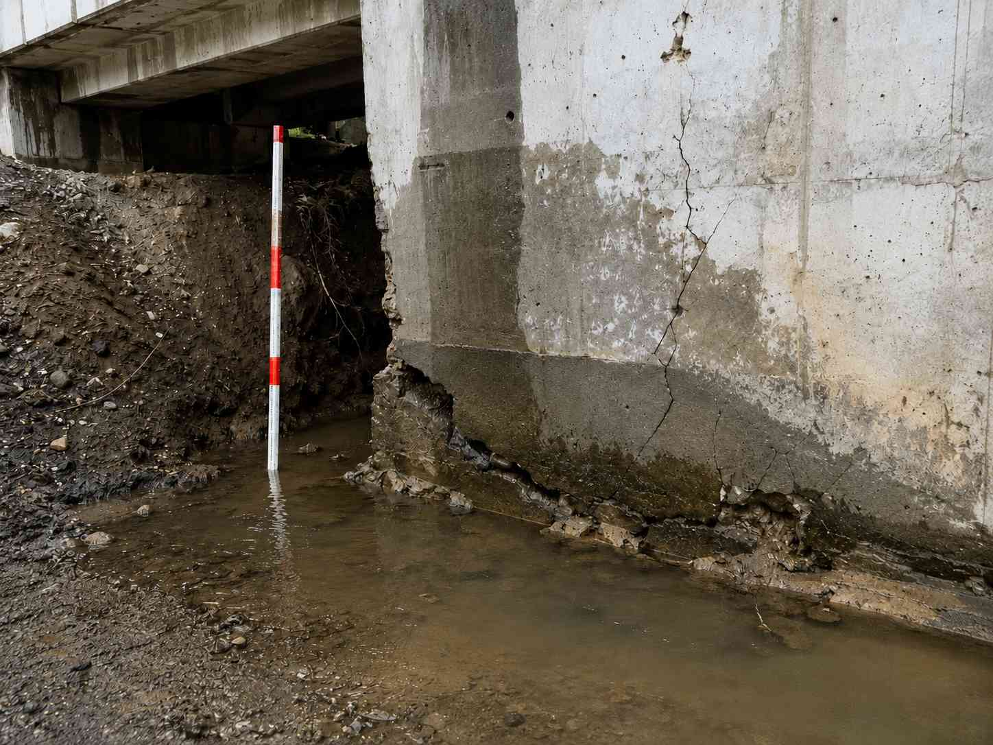 Water intrusion and soil erosion beneath a concrete bridge foundation with pooling water, cracks, and moisture stains.